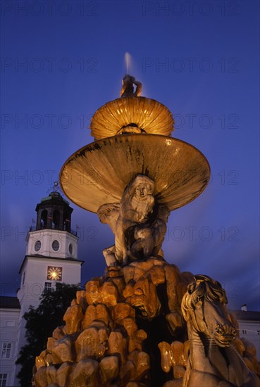 AUSTRIA, Salzburg, Part view of Baroque fountain with statues of horse and contorted figures and the Glockenspiel Carillon tower behind at night.