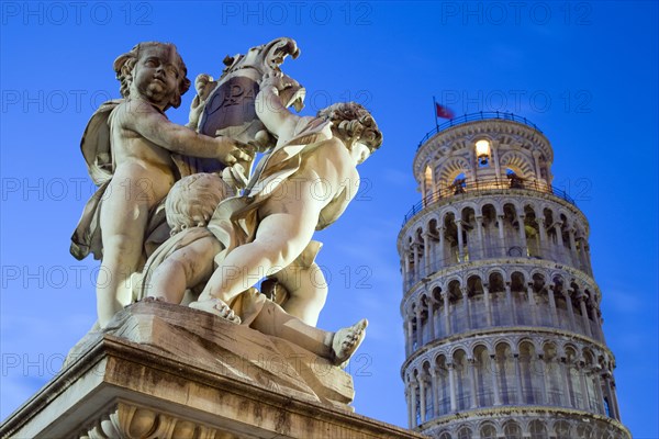 ITALY, Tuscany, Pisa, An illuminated statue in The Piazza del Duomo of cherubs holding a shield bearing the Cross of Pisa with the Leaning Tower beyond all within the UNESCO World Heritage site