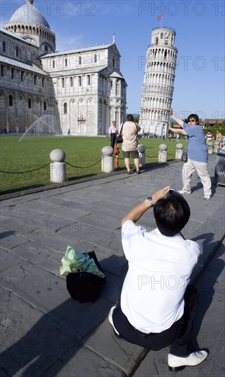 ITALY, Tuscany, Pisa, Asian male tourist taking a photograph of another Asian man pretending to hold up the Leaning Tower of Pisa in The Campo dei Miracoli or Field of Miracle all within the UNESCO World Heritage site
