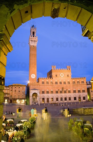 ITALY, Tuscany, Siena, "Early evening in the Piazza del Campo seen through an archway with people at restaurant tables and walking in the square. The illuminated Torre del Mangia, the second tallest belltower in Italy, is beside the Palazzo Publico at the far side of the piazza"
