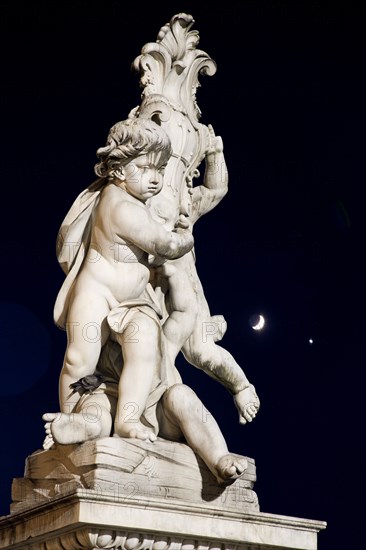 ITALY, Tuscany, Pisa, An illuminated statue in The Piazza del Duomo of cherubs holding a shield bearing the Cross of Pisa with a half moon shining in the night sky beyond