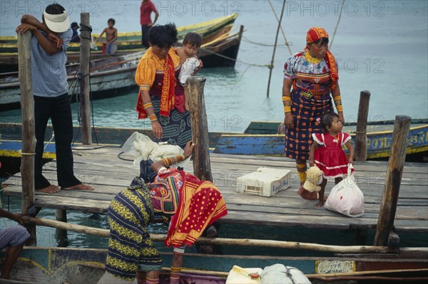 PANAMA, San Blas Islands Tikantiki , Kuna Indians, Kuna Indian women wearing brightly coloured traditional mola panel blouses or dulemolas arriving by canoe at island jetty with children. Cuna Caribbean Niadup American Central America Colored Female Woman Girl Lady Hispanic Kids Latin America Latino Panamanian West Indies