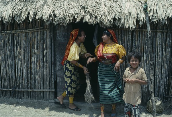 PANAMA, San Blas Islands Tikantiki , Kuna Indians, Kuna Indian women wearing brightly coloured traditional mola panel blouses or dulemolas joking outside their home with young boy standing at side. Cuna Caribbean American Central Central America Colored Female Woman Girl Lady Hispanic Kids Latin America Latino Panamanian West Indies