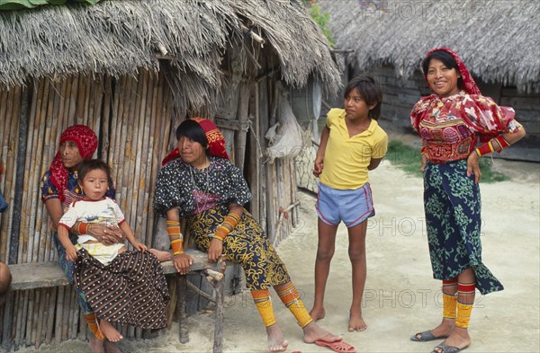 PANAMA, San Blas Islands, Kuna Indians, Kuna Indian family with women wearing brightly coloured tradtional mola panel blouses or dulemolas outside their thatched home  their legs bound with traditional design strings of coloured glass beads Cuna Caribbean  house American Central America Colored Female Woman Girl Lady Hispanic Kids Latin America Latino Panamanian West Indies