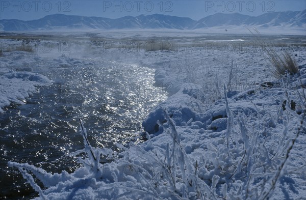MONGOLIA, Mongolian Steppe, Mid-winter at Bigersum negdel collective. River runs through desert snow-covered cultivated land with steam rising off water at around -25.C. Altai mountains in background.