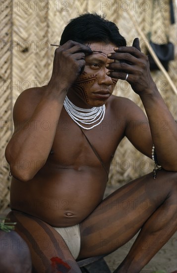 COLOMBIA, North West Amazon, Tukano Indigenous People, "Barasana man looking in mirror to apply red Achiote facial paint,  thick dark purple we leaf juice vertical stripes painted on legs  wearing multi stranded white glass bead necklace.  Tukano sedentary Indian tribe North Western Amazonia body decoration American Colombian Columbia Hispanic Indegent Latin America Latino Male Men Guy South America Tukano  "