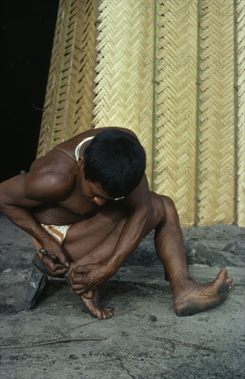 COLOMBIA, North West Amazon, Tukano Indigenous People, Barasana man removing palm spine from foot outside entrance to maloca/communal home. Tukano sedentary Indian tribe North Western Amazonia American Colombian Columbia Hispanic Indegent Latin America Latino Male Men Guy South America Tukano Male Man Guy One individual Solo Lone Solitary