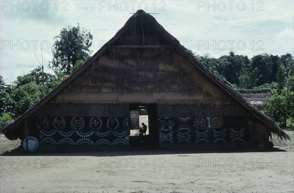 COLOMBIA, North West Amazon, Tukano Indigenous People, Makuna communal home or maloca with white and yellow clay and  charcoal painted design on exterior facade.  Man basket-making silhouetted in rear women's entrance. Tukano  Makuna Indian North Western Amazonia maloca American Colombian Columbia Hispanic Indegent Latin America Latino Male Men Guy South America Tukano