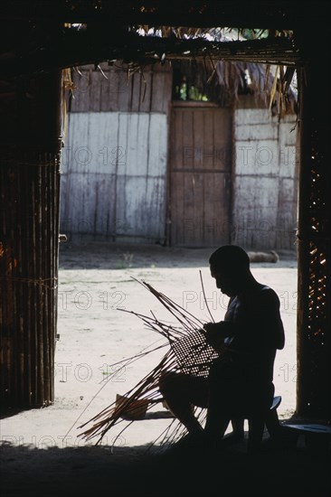 COLOMBIA, North West Amazon, Tukano Indigenous People, Makuna head man Ignacio making cane basket silhouetted in entrance to maloca or communal home. Tukano  Makuna Indian North Western Amazonia moloka American Colombian Columbia Hispanic Indegent Latin America Latino Male Men Guy South America Tukano Male Man Guy