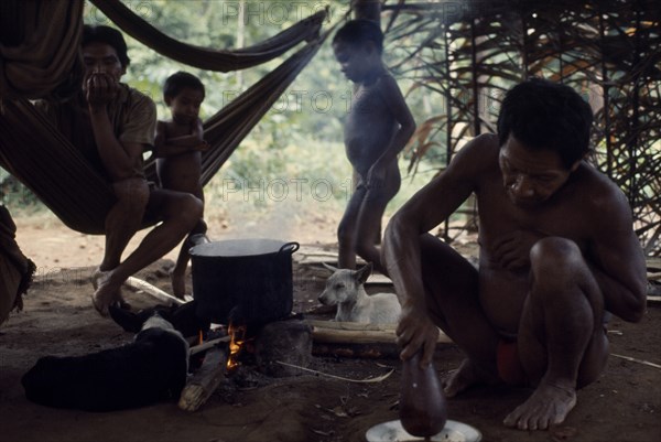 COLOMBIA, North West Amazon, Vaupes, Maku Indian men and children inside palm-thatched shelter/home with hunting dogs lying beside open fire and cooking pot. indigenous tribe indian nomadic American Colombian Columbia Hispanic Indegent Kids Latin America Latino Male Man Guy South America Vaupes Male Men Guy Vaupes