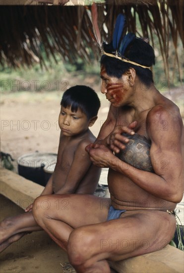 COLOMBIA, North West Amazon, Vaupes, Maku hunter  face painted with red Achiote marks and wearing  macaw feather crown in preparation for dance  sits beside young boy. indigenous tribe indian nomadic American Colombian Columbia Hispanic Indegent Kids Latin America Latino Male Men Guy South America Vaupes Immature Male Man Guy Vaupes