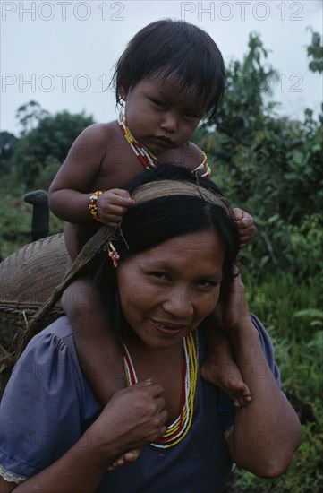 COLOMBIA, North West Amazon, Tukano Indigenous People, Makuna mother carrying her baby son on her shoulders and basket on her back supported by head strap.  Both  wearing necklaces of white red and yellow glass beads. Tukano  Makuna Indian North Western Amazonia family American Babies Colombian Columbia Hispanic Indegent Kids Latin America Latino Mum South America Tukano