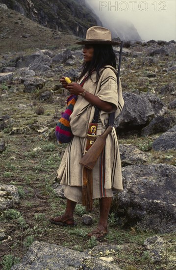 COLOMBIA, Sierra Nevada de Santa Marta, Ika, Ika shepherd in the high Sierra carrying rifle on his shoulder  takes lime from his small poporo gourd to place in the wad of coca leaves in his cheek. Lime is the catalyst which releases a very small amount of the cocaine alkaloid from coca leaves. Coca keeps away the pangs of hunger & cold  Arhuaco Aruaco indigenous tribe American Colombian Colombia Hispanic Indegent Latin America Latino South America  Arhuaco Aruaco indigenous tribe American Colombian Columbia Hispanic Indegent Latin America Latino South America Farming Agraian Agricultural Growing Husbandry  Land Producing Raising Immature One individual Solo Lone Solitary 1 Agriculture Single unitary Young Unripe Unripened Green