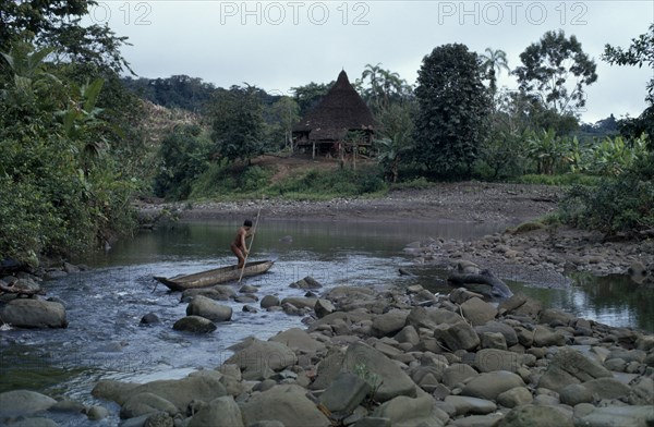 COLOMBIA, Choco, Embera Indigenous People, "Embera man using pole to steer wooden dug out canoe along river, approaches the port of his stilted and thatched family home set back amongst cleared trees and vegetation. Pacific coastal region boat piragua tribe "