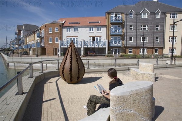 ENGLAND, West Sussex, Shoreham-by-Sea, Ropetackle housing development on the banks of the river Adur. Young woman reading a book on the riverbank promenade. A regenerated brownfield former industrial area.