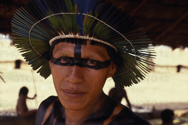 BRAZIL, Mato Grosso, Indigenous Park of the Xingu, Head and shoulders portrait of young male Panara leader wearing ceremonial crown made from green/blue parrot feathers and black face and body paint. Formally known as Kreen-Akrore  Krenhakarore  Krenakore  Krenakarore  Amazon American Brasil Brazilian Indegent Kreen Akore Latin America Latino South America  Formally known as Kreen-Akrore  Krenhakarore  Krenakore  Krenakarore  Amazon American Brasil Brazilian Indegent Kreen Akore Latin America Latino South America Immature One individual Solo Lone Solitary 1 Single unitary Young Unripe Unripened Green