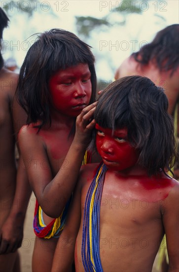 BRAZIL, Mato Grosso, Indigenous Park of the Xingu, Young Panara girls applying red karajuru face and body paint in preparation for dance.  Both wearing multiple strands of blue red and yellow beads across upper body. Formally known as Kreen-Akrore  Krenhakarore  Krenakore  Krenakarore  Amazon American Brasil Brazilian Indegent Kids Kreen Akore Latin America Latino South America Immature
