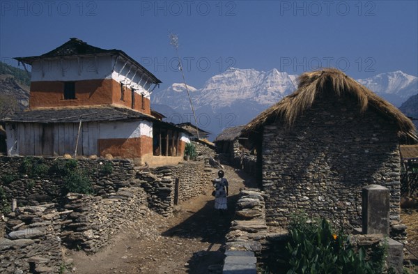 NEPAL, Dhorpatan Trek, Dhorpatan, Woman walking between stone houses in Dharapani Village and the Dhaulagiri Himal snow capped mountain behind in the distance