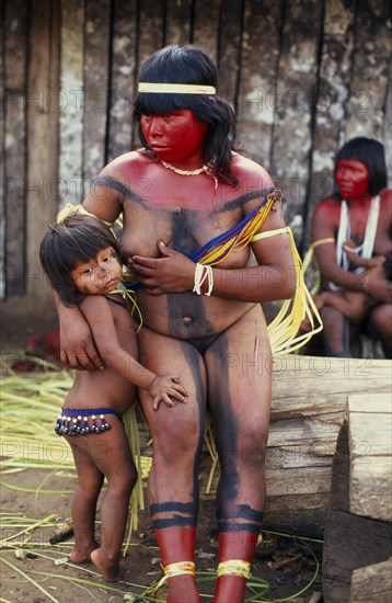 BRAZIL, Mato Grosso, Indigenous Park of the Xingu, Panara woman breastfeeding daughter before dance  with face and neck painted with red karajuru and wearing multi-strand of beads across upper body and long strips of plant fibres tied around upper arms.  Formally known as Kreen-Akrore  Krenhakarore  Krenakore  Krenakarore  Amazon American Brasil Brazilian Female Women Girl Lady Indegent Kreen Akore Latin America Latino South America Female Woman Girl Lady Immature Kids