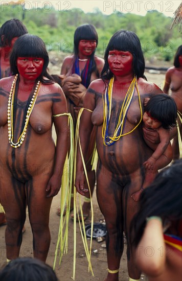 BRAZIL, Mato Grosso, Indigenous Park of the Xingu, Panara women painted with red karajuru and wearing multi-strands of beads across upper body and long strips of plant fibres tied around upper arms in preparation for dance.  Formally known as Kreen-Akrore  Krenhakarore  Krenakore  Krenakarore  Amazon American Brasil Brazilian Female Women Girl Lady Indegent Kreen Akore Latin America Latino South America Female Woman Girl Lady Immature