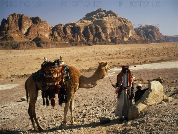 JORDAN, People, "Bedouin man and child standing beside camel carrying decorated, woven saddle and panniers in desert area near Wadi Rum."
