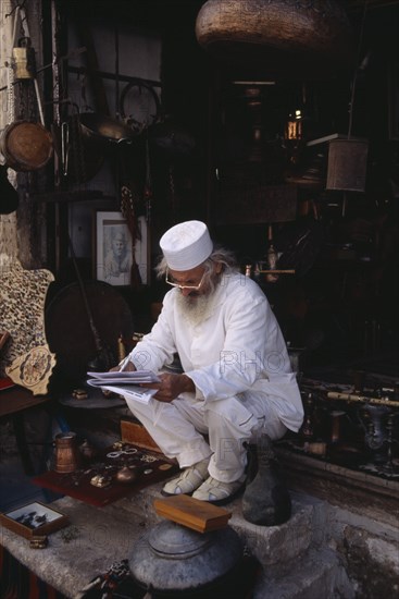 BOSNIA & HERZEGOVINA, Balkan Peninsula, Mostar, Elderly man wearing white sitting on step at entrance to antique shop to read papers.