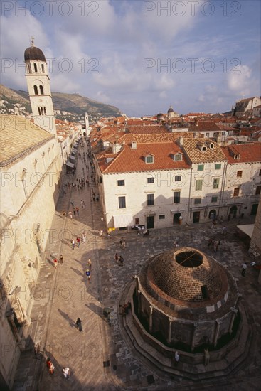 CROATIA, Dalmatia, Dubrovnik, "Elevated view over cobbled square with circular stone building in centre, white painted houses with red tiled rooftops, people and bell tower on left."