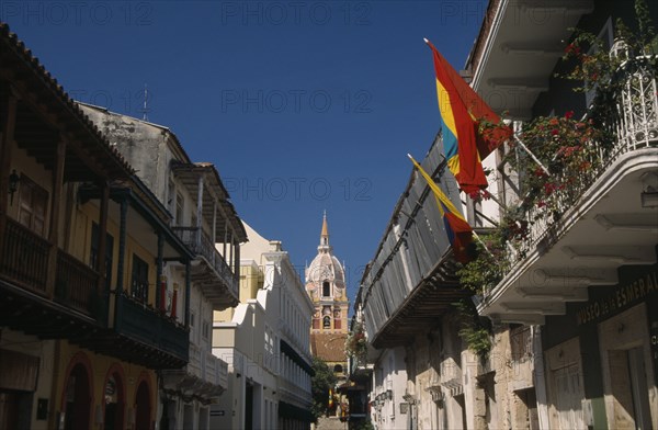 COLOMBIA, Bolivar, Cartagena, "View along Calle San Pedro Claver towards cathedral bell tower and spire.  Narrow street lined with buildings with upper balconies, flags flying in foreground."