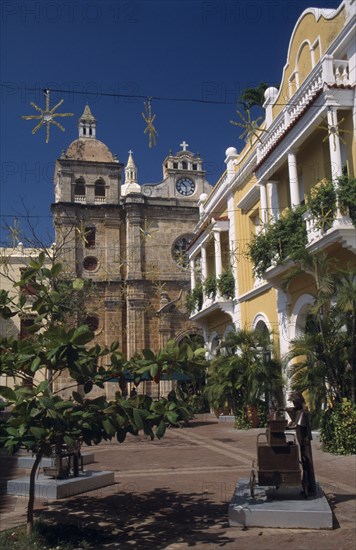 COLOMBIA, Bolivar, Cartagena, Plaza de San Pedro Claver.  Yellow and white colonial facade of building on right with part view of Church of San Pedro Claver beyond.  Sculpture and trees in foreground.