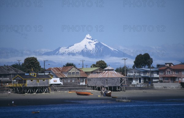 CHILE, Los Lagos, Architecture, Coastal village buildings near Chiloe from sea with snow capped volcanic peak wreathed in cloud behind.