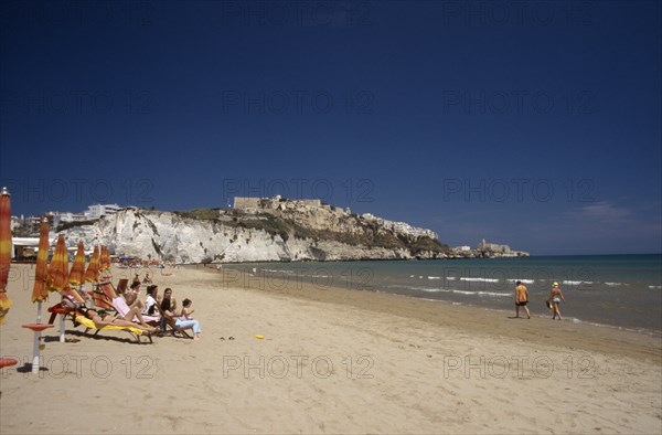 ITALY, Puglia, Foggia, Vieste.  Line of sunbathers on stretch of sandy beach overlooked by castle and apartments on white cliffs beyond.