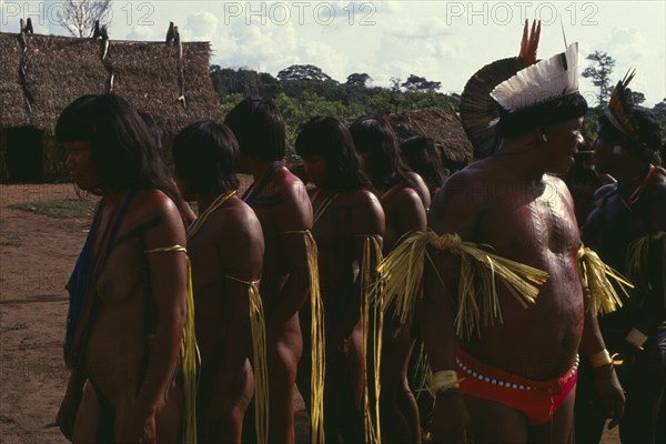 BRAZIL, Mato Grosso, Indigenous Park of the Xingu, Panara men wearing crowns or head-dresses of feathers beside women painted with red karajuru and wearing colourful strands of beads and with long strips of plant fibres tied around upper arms during dance. Formerly known as Kreen-Akrore  Krenhakarore  Krenakore  Krenakarore  Amazon American Brasil Brazilian Colorful Female Woman Girl Lady Indegent Kreen Akore Latin America Latino Male Man Guy South America Female Women Girl Lady Male Men Guy