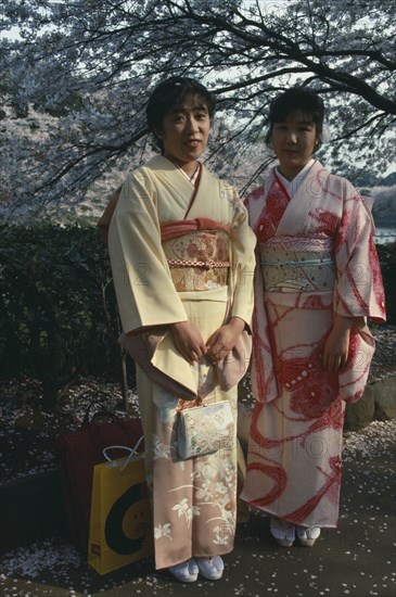 JAPAN, Honshu, Tokyo, Two young women wearing traditional kimonos whilst stood under a cherry blossom tree.