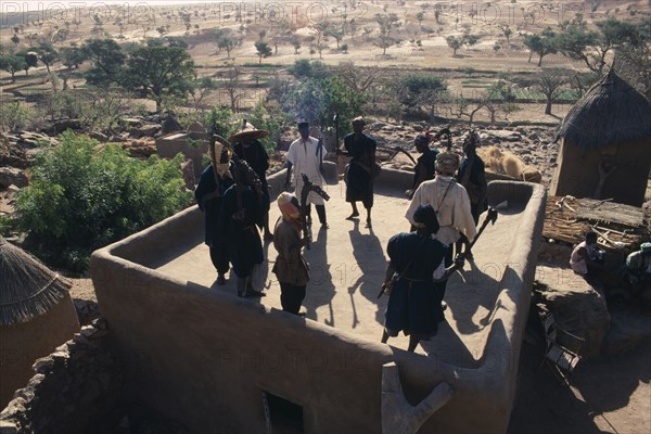 20086075 MALI People Dogon Members of ancient Dogon society performing rooftop dance holding carved staffs with view over surrounding village and landscape.