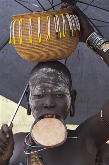 ETHIOPIA, South Omo Valley, Mursi Tribe, Woman with lip plate holding umbrella to shelter from sun with basket on head