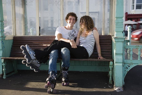 ENGLAND, East Sussex, Brighton, "Young couple sat on bench on the seafront promenade. One wearing Rollerblades, one wearing Rollerboots"