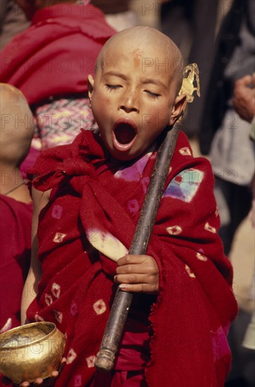 NEPAL, Kathmandu Valley, Bhaktapur, Young monk yawning during a ceremony and wearing red robes outside a small monastery in the back streets