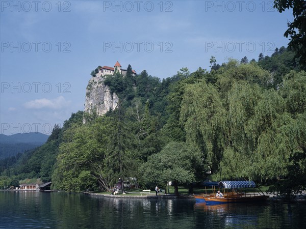 SLOVENIA, Lake Bled, Tourist boats on Lake Bled with castle partly seen amongst trees high above.  Few people on lake shore below.