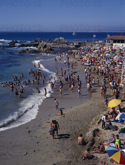 CHILE, Valparaiso Region, Los Lilenes, Crowded beach near Vina del Mar.  People sunbathing and playing in the surf.  Rocks and distant yacht beyond.
