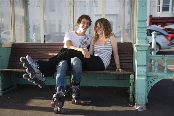 ENGLAND, East Sussex, Brighton, "Young couple sat on bench on the seafront promenade. One wearing Rollerblades, one wearing Rollerboots"