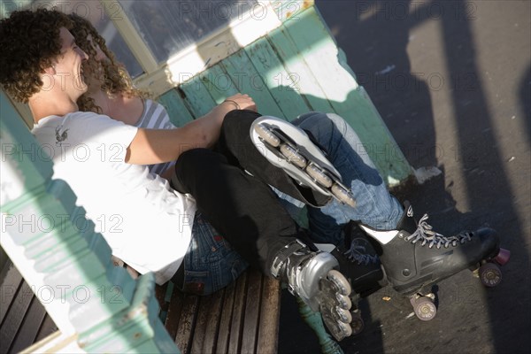 ENGLAND, East Sussex, Brighton, "Young couple sat on bench on the seafront promenade. One wearing Rollerblades, one wearing Rollerboots"