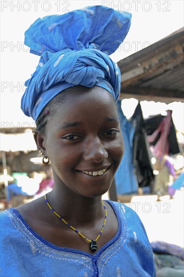 GAMBIA, Western Gambia, Tanji, Tanji Village.  Head and shoulders portrait of smiling African girl with a gold tooth and wearing blue dress and traditional tied head scarf in bright sunshine.