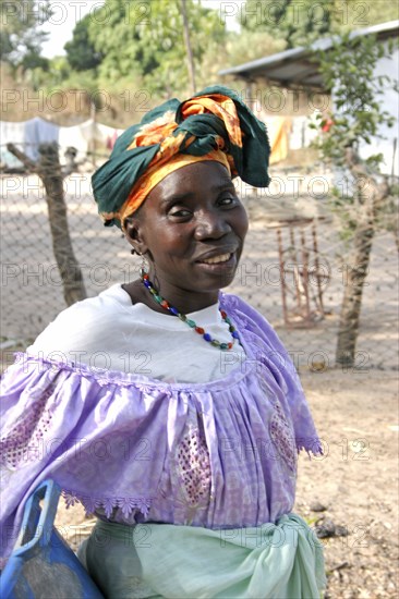 GAMBIA, Western Gambia, Tanji, Tanji village.  Three-quarter portrait of woman wearing traditional head tie or scarf and bead necklace posing in front of her home compound.