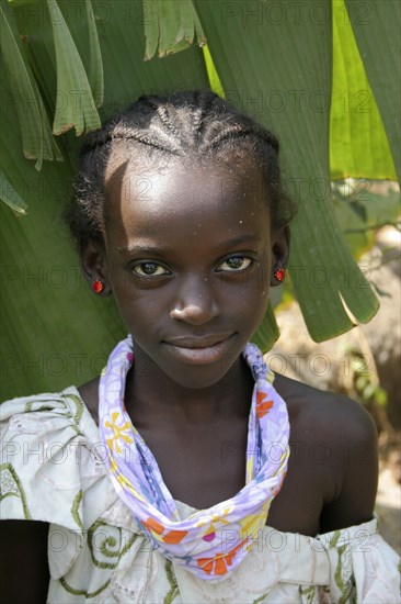 GAMBIA, Western Gambia, Tanji, Tanji village.  Head and shoulders portrait of smiling young girl with braided hair and almond shaped eyes wearing patterned scarf tied around her neck and red earrings.