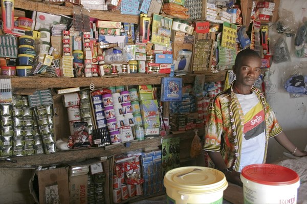 GAMBIA, Western Gambia, Tanji, "Tanji Village.  Young, male owner of a grocery shop standing behind wooden counter in front of shelves with display of various products. "
