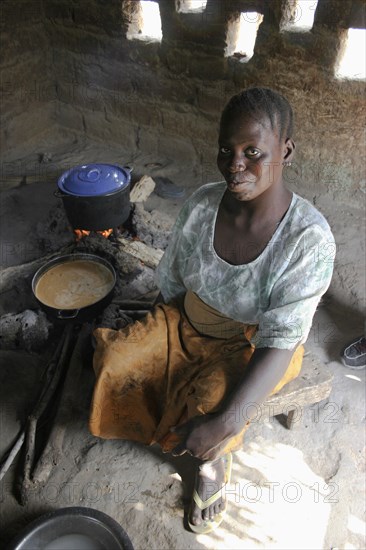 GAMBIA, Western Gambia, Tanji, "Tanji Village.  Smiling woman cooking traditional dish of groundnut sauce called mafay over open fire in kitchen which is outside of the house.  Sunlight shining through small, square openings in wall behind."