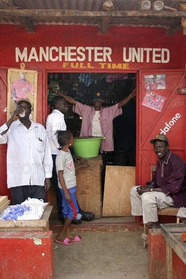 GAMBIA, Atlantic Coast, Banjul, "Shop in Gambia's capital Banjul painted red and white, the colours of the Manchester United football strip and decorated with slogans and posters with group of African supporters of the team gathered outside and in doorway."