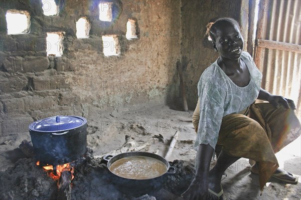 GAMBIA, Western Gambia, Tanji, "Young woman cooking a traditional Gambian dish of groundnut sauce called mafay over open fire in kitchen situated outside  house with sunlight filtering through small, square openings in the wall behind."