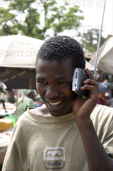 GAMBIA, Atlantic Coast, Banjul, "Albert Market, Russell Street.  Young African man smiling  while listening to a radio which he is holding next to his ear."