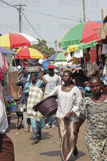 GAMBIA, Western Gambia, Serekunda, "Bakau Market, Atlantic Road.  Busy market scene with crowds of people, and roadside stalls beneath colourful striped sun umbrellas. "